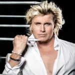 Hans Klok poseert in een open wit shirt tussen scherpe metalen staven tegen een zwarte achtergrond.