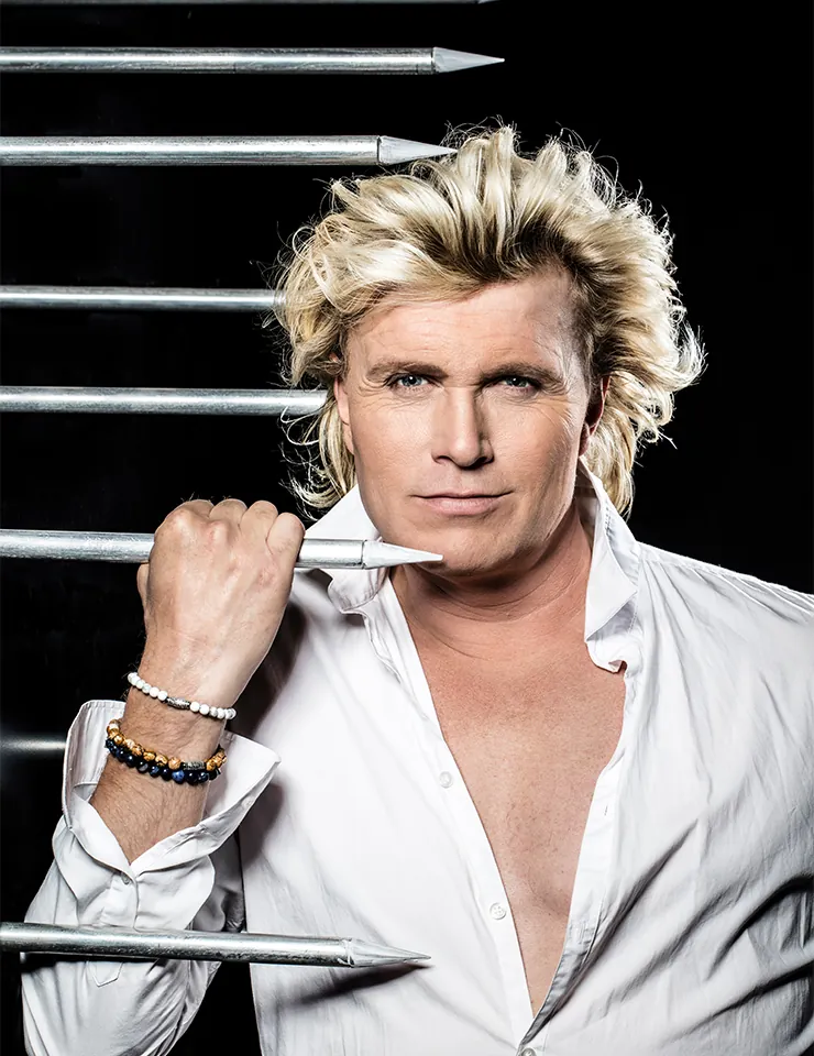 Hans Klok poseert in een open wit shirt tussen scherpe metalen staven tegen een zwarte achtergrond.