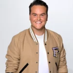 Henk Dissel met kort donker haar en een beige varsity-jack poseert lachend voor een neutrale achtergrond.