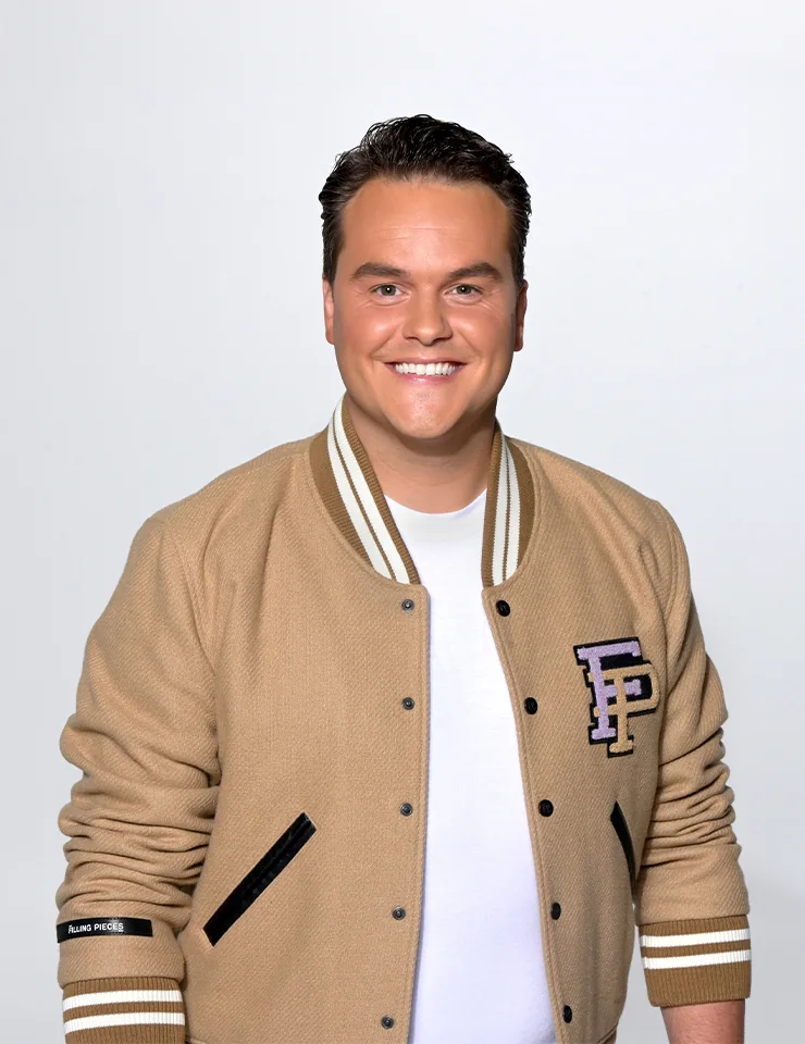 Henk Dissel met kort donker haar en een beige varsity-jack poseert lachend voor een neutrale achtergrond.