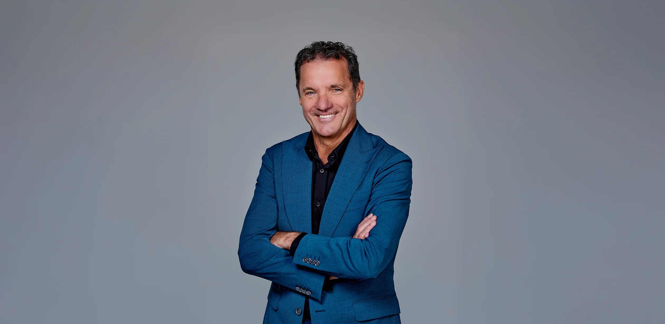 John de Bever in blauw pak poseert glimlachend met armen over elkaar tegen een grijze achtergrond.