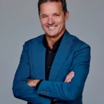 John de Bever in blauw pak met zwarte overhemd poseert glimlachend met gekruiste armen tegen een grijze achtergrond.