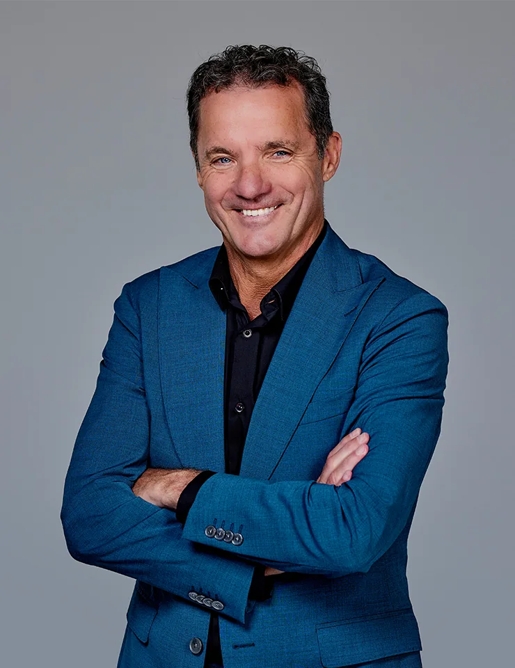 John de Bever in blauw pak met zwarte overhemd poseert glimlachend met gekruiste armen tegen een grijze achtergrond.