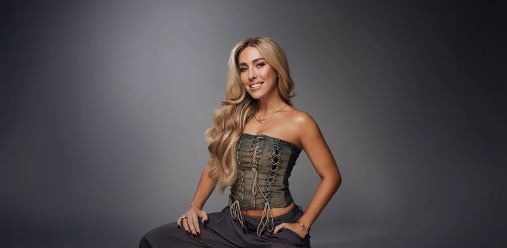 Portret van Kimberly Fransens met lang blond haar, gekleed in een korset-top en zittend voor een grijze achtergrond.