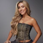 Kimberly Fransens met lang blond haar poseert in een denim korset-top en donkere broek tegen een grijze achtergrond.