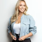 Monique Smit met lang blond haar poseert lachend in een lichtblauw jasje en witte top tegen een witte achtergrond.