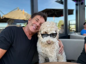 Gerard Joling poseert lachend met zijn hond Buddy, die een zonnebril draagt.