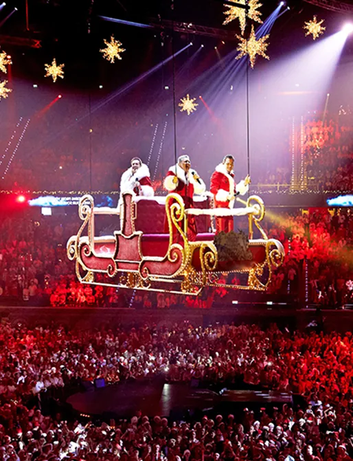 De Toppers in kerstkostuum zweven in een verlichte arrenslee boven een feestelijk publiek tijdens de Kersttoppers-show.