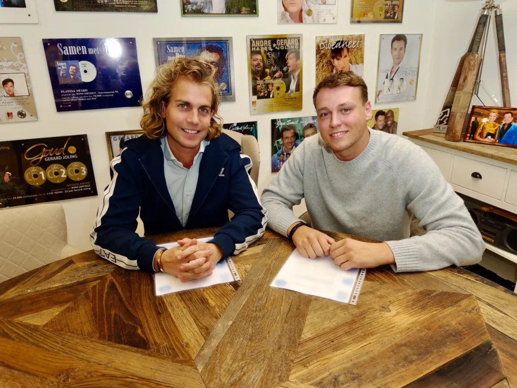 Ramon Fortier en Glenn zitten samen aan een houten tafel met documenten voor zich, voor een muur vol muziek- en artiestenposters.