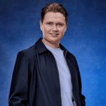 Portret van Kelsey Verbrugge tegen een blauwe achtergrond, gekleed in een donker jack en licht T-shirt.