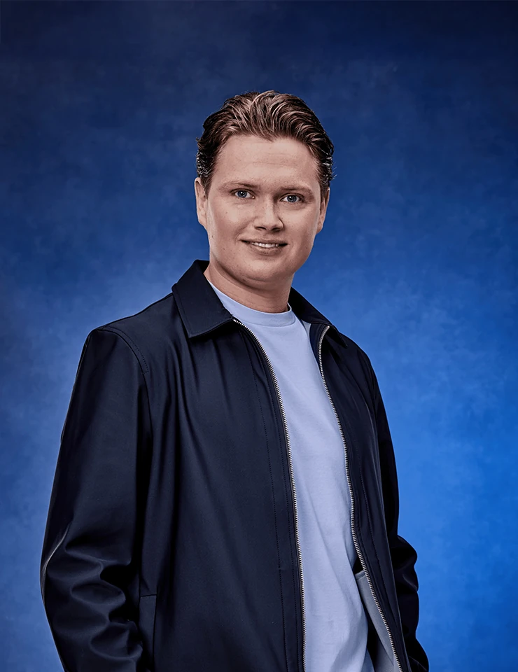 Portret van Kelsey Verbrugge tegen een blauwe achtergrond, gekleed in een donker jack en licht T-shirt.