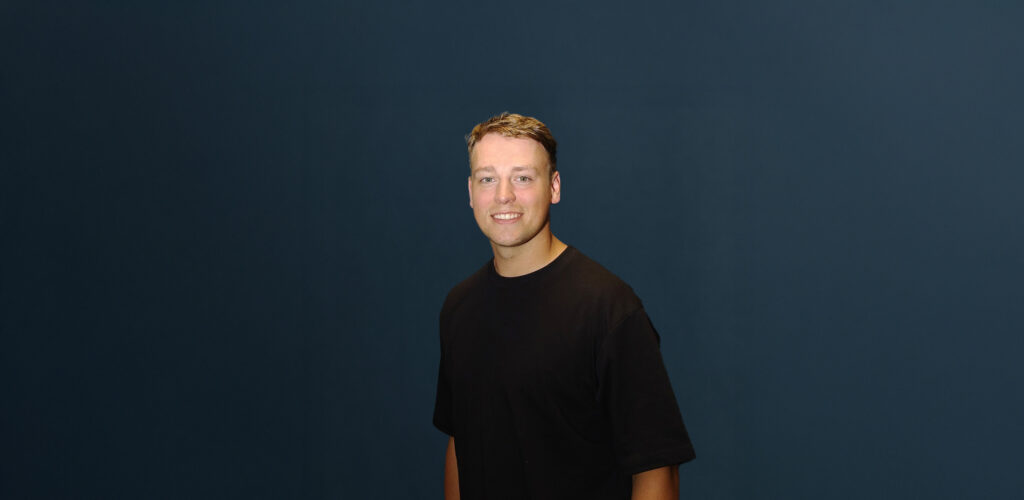 Ramon Fortier met kort bruin haar en zwart T-shirt poseert voor een effen donkerblauwe achtergrond.