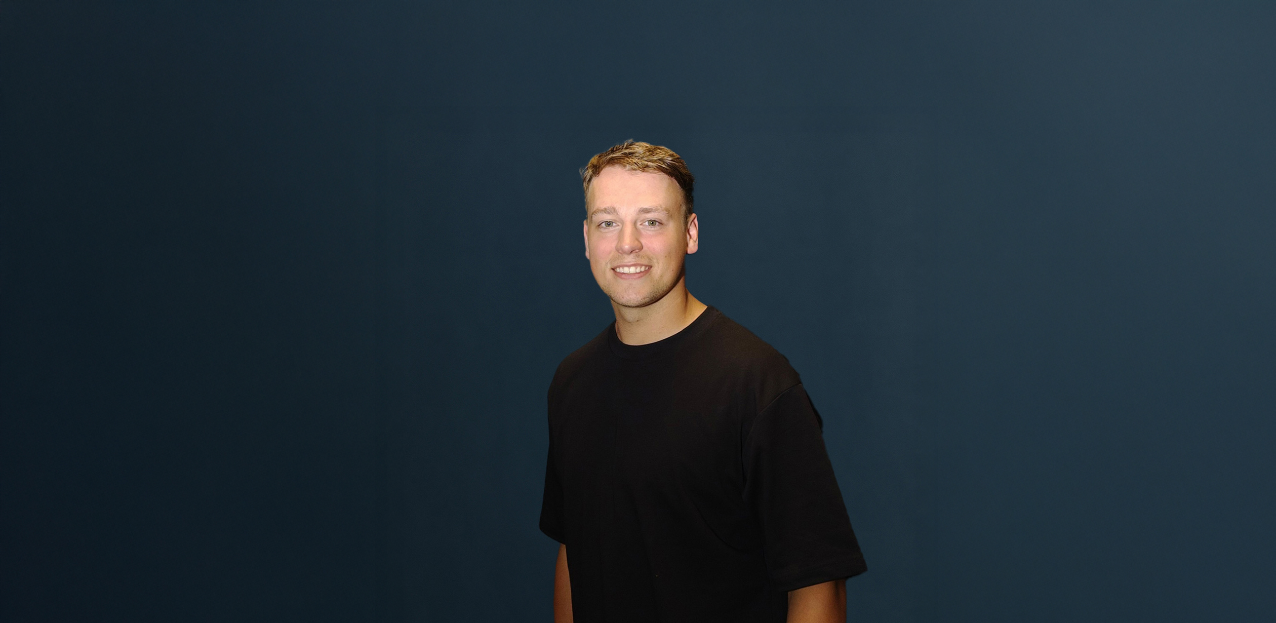 Ramon Fortier met kort bruin haar en zwart T-shirt poseert voor een effen donkerblauwe achtergrond.