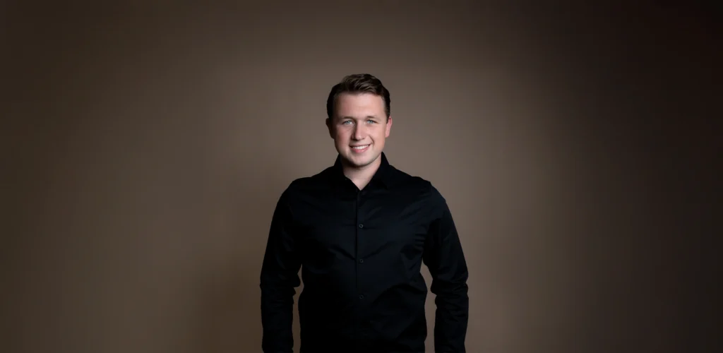 Teun Landzaat poseert lachend voor een neutrale, bruine achtergrond in een zwarte blouse.