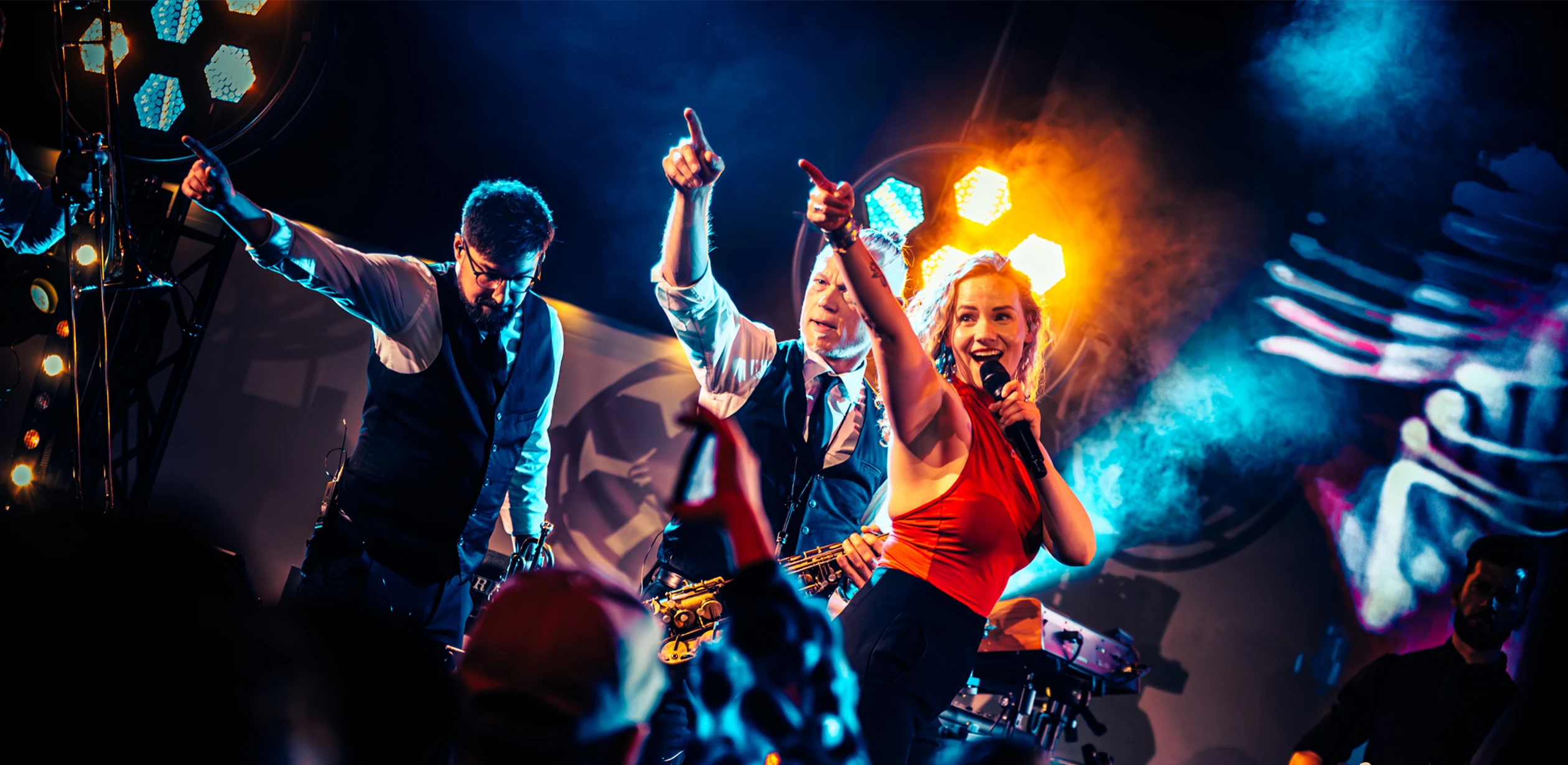 Zangeres en blazers van de band Boston Tea Party treden energiek op op een podium met kleurrijke verlichting.