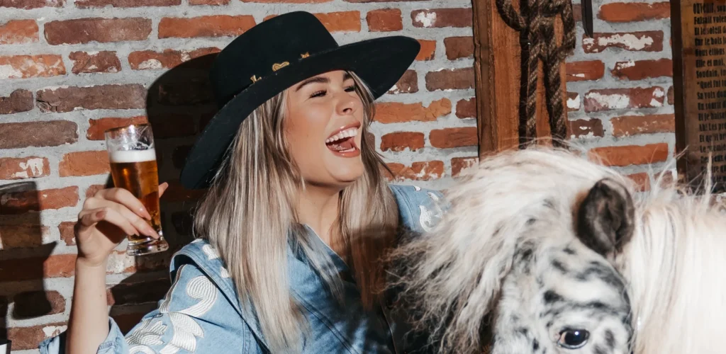 Jorieke Sterken lacht terwijl ze een glas bier vasthoudt en naast een gevlekt paard zit.