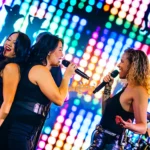 Drie zangeressen van de band Boston Tea Party zingen op het podium voor een kleurrijk verlicht scherm.