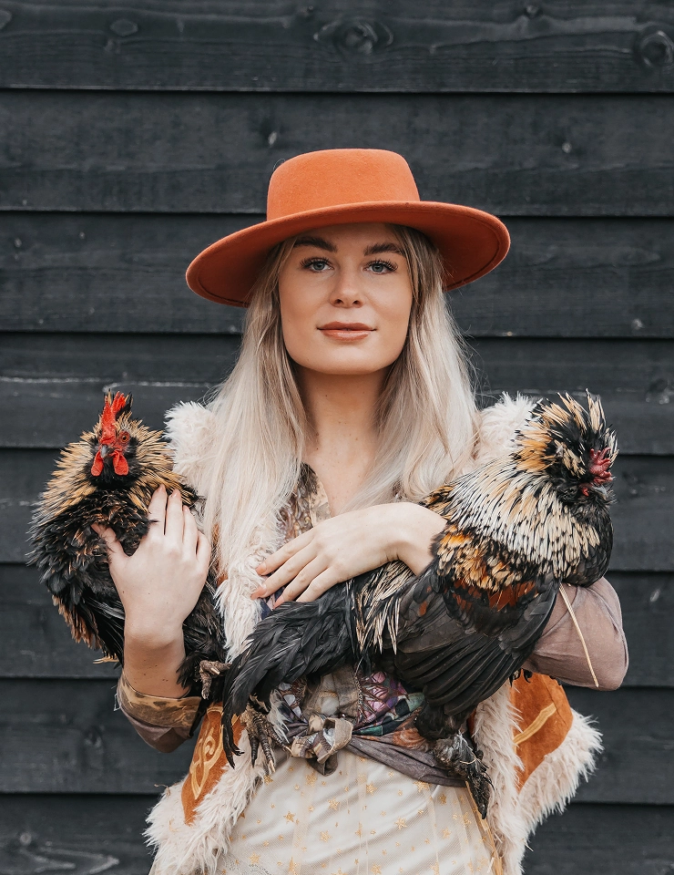 Jorieke Sterken met lange blonde haren en oranje hoed houdt twee kleurrijke kippen vast voor een donkere houten achtergrond.