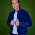 Quincy in blauwe jas en jeans poseert lachend voor een groene achtergrond.