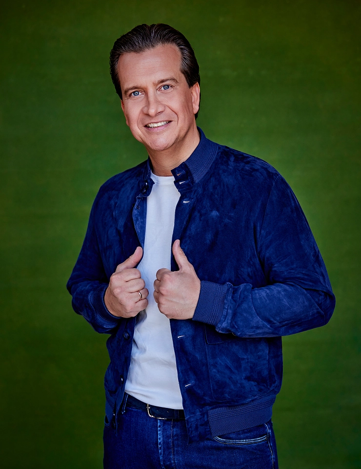 Quincy in blauwe jas en jeans poseert lachend voor een groene achtergrond.