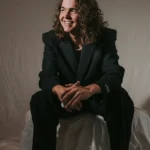 Sem Rozendaal zit lachend op een kruk, gekleed in een donker pak tegen een neutrale achtergrond.