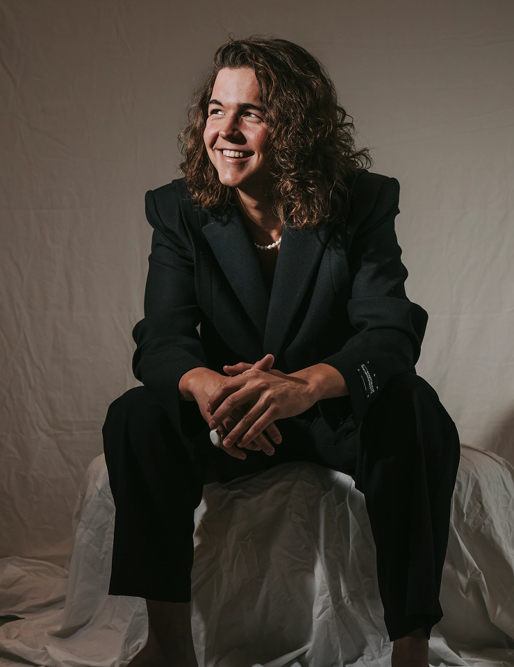 Sem Rozendaal zit lachend op een kruk, gekleed in een donker pak tegen een neutrale achtergrond.