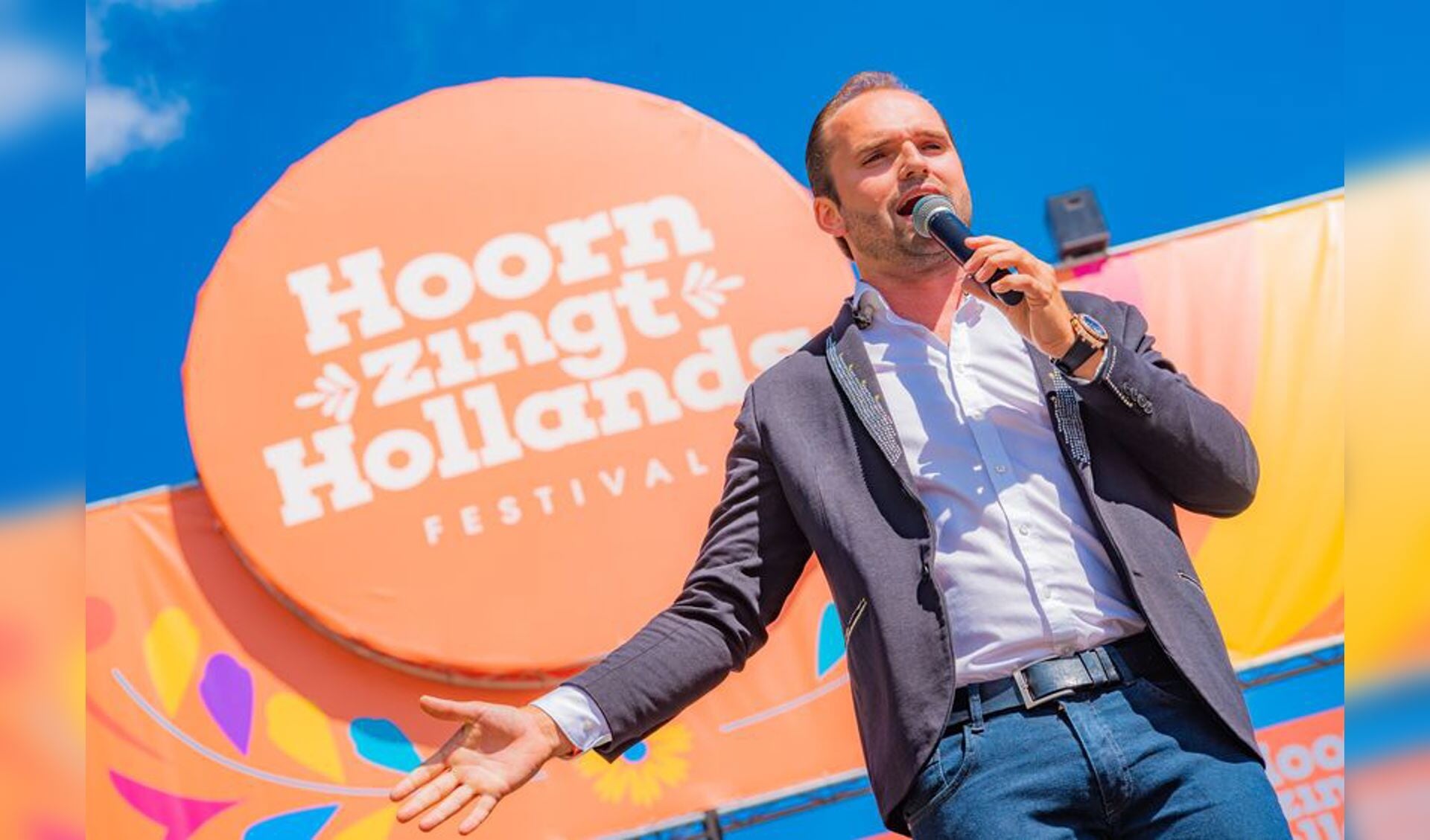 Bryan Stricker zingt op het podium tijdens Hoorn Zingt Hollands, met een microfoon in de hand voor een kleurrijke festivalaankleding.