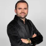 Bryan Stricker met kort haar en een zwarte blouse met colbert poseert lachend voor een neutrale witte achtergrond.