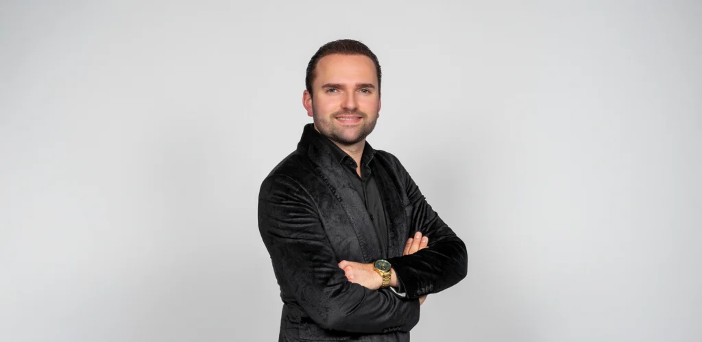 Portretfoto Bryan Stricker met kort haar en een zwarte blouse met colbert voor een witte achtergrond.