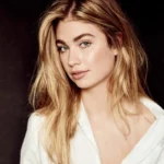 Portret van Roxy Dekker met los blond haar en een witte blouse tegen een donkere achtergrond.