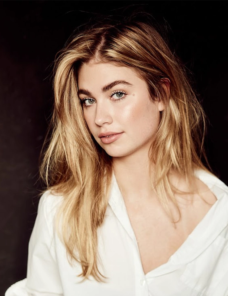 Portret van Roxy Dekker met los blond haar en een witte blouse tegen een donkere achtergrond.